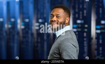 Young Black Data Center IT Technician Standing in Server Rack Corridor and Smiling to the Camera. Stock Photo