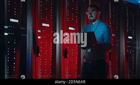 Angry Bearded IT Specialist in Glasses is Walking Through Server Rack Corridor in Data Center and Holding Laptop. Doing Emergency Maintenance Work Stock Photo