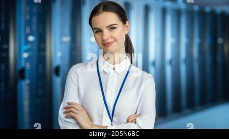 Female IT Specialist is standing at the Camera in Data Center Next to Server Racks and Looking at the Camera. Her Arms Crossed and She Smiles to the Stock Photo