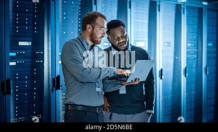 Bearded IT Technician in Glasses with Laptop Computer and Black Male Engineer Colleague are Using Laptop in Data Center while Working Next to Server Stock Photo