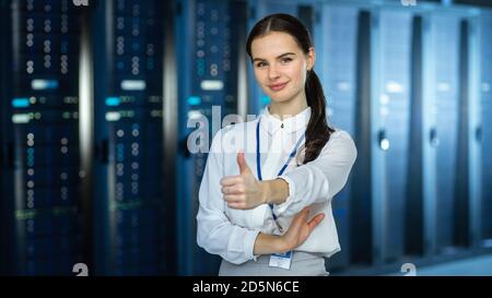 Female IT Specialist is standing at the Camera in Data Center Next to Server Racks and Looking at the Camera. She Shows Thumb up and Smiles to the Stock Photo