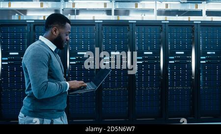Black Data Center IT Technician Walking Through Server Rack Corridor with a Laptop Computer. He is Visually Inspecting Working Server Cabinets. Stock Photo