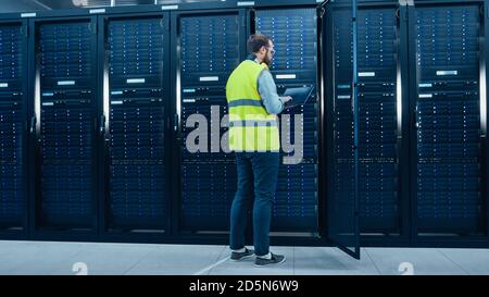 IT Engineer in High Visibility Vest is Working on Laptop in Data Center while Standing Before Server Rack. Running Diagnostics or Doing Maintenance Stock Photo