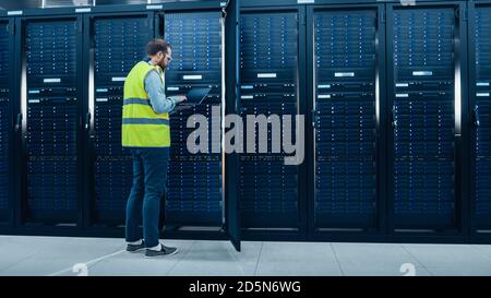 IT Engineer in High Visibility Vest is Working on Laptop in Data Center while Standing Before Server Rack. Running Diagnostics or Doing Maintenance Stock Photo