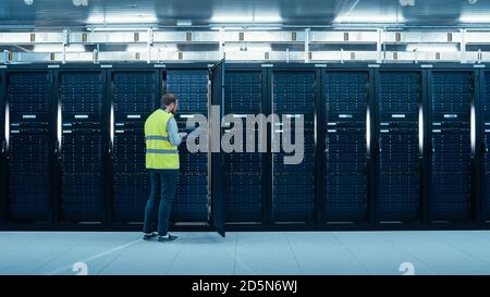 IT Engineer in High Visibility Vest is Working on Laptop in Data Center while Standing Before Server Rack. Running Diagnostics or Doing Maintenance Stock Photo