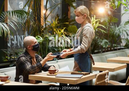 Man in protective mask using his mobile phone to pay for his order to the waiter during pandemic at the restaurant Stock Photo