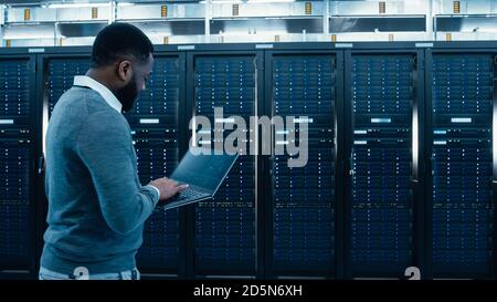 Black Data Center IT Technician Walking Through Server Rack Corridor with a Laptop Computer. He is Visually Inspecting Working Server Cabinets. Stock Photo