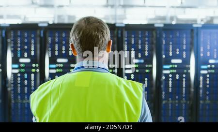 Back Shot of an IT Administrator in High Visibility Vest Walking Towards a Server Rack in Data Center Room. Stock Photo