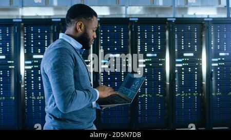 Black Data Center IT Technician Walking Through Server Rack Corridor with a Laptop Computer. He is Visually Inspecting Working Server Cabinets. Stock Photo