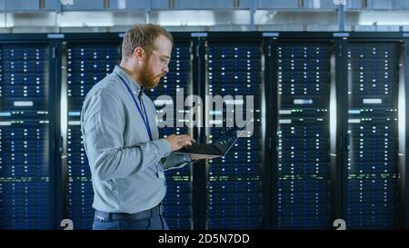 Bearded Data Center IT Engineer in Glasses is Walking Through Server Rack Corridor with a Laptop Computer. He is Visually Inspecting Working Server Stock Photo