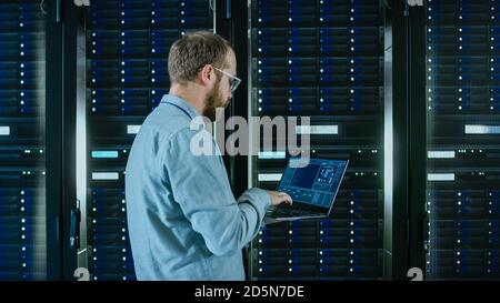 Bearded Data Center IT Engineer in Glasses is Walking Through Server Rack Corridor with a Laptop Computer. He is Visually Inspecting Working Server Stock Photo