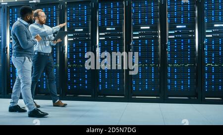 IT Technician with a Laptop Computer and Black Male Engineer Colleague are Talking in Data Center while Walking Next to Server Racks. Running Stock Photo