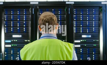 Back Shot of an IT Administrator in High Visibility Vest Walking Towards a Server Rack in Data Center Room. Stock Photo