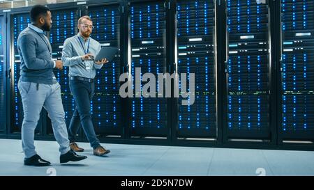 IT Technician with a Laptop Computer and Black Male Engineer Colleague are Talking in Data Center while Walking Next to Server Racks. Running Stock Photo