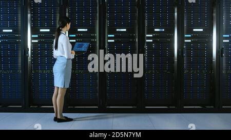 Beautiful Data Center Female IT Technician Walking Through Server Rack Corridor with a Laptop Computer. She Stops and Visually Inspects Working Server Stock Photo