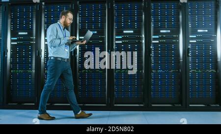 Bearded Data Center IT Professional Walking Through Server Rack Corridor with a Laptop Computer. He Stops and Wirelessly Inspects Working Server Stock Photo