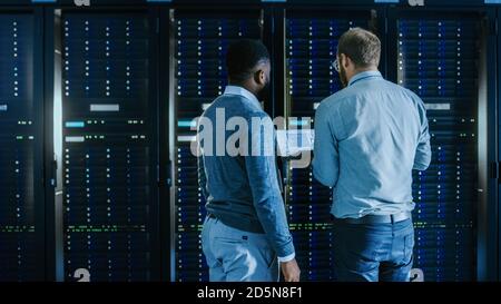Bearded IT Technician in Glasses with a Laptop Computer and Black Male Engineer Colleague are Working in Data Center while Working Next to Server Stock Photo