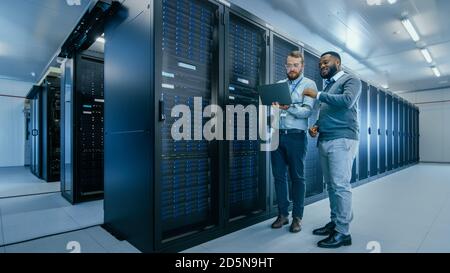 Bearded IT Technician in Glasses with a Laptop Computer and Black Male Engineer Colleague are Talking in Data Center while Working Next to Server Stock Photo