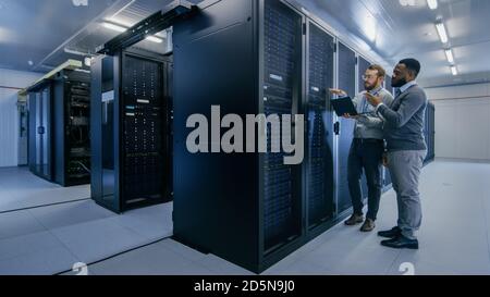Bearded IT Technician in Glasses with a Laptop Computer and Black Male Engineer Colleague are Talking in Data Center while Working Next to Server Stock Photo