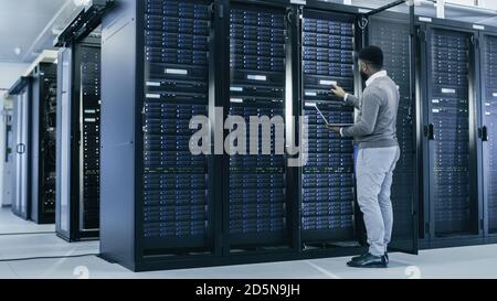 Black IT Specialist is Working on Laptop in Data Center while Standing Before Server Rack. Running Diagnostics, Putting in Data or Doing Maintenance Stock Photo