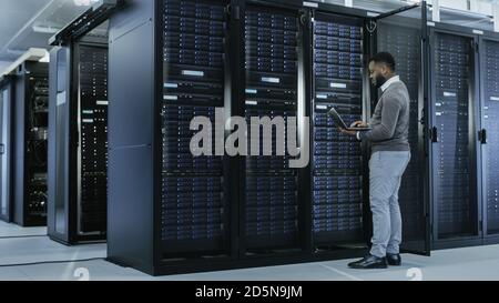 Black IT Specialist is Working on Laptop in Data Center while Standing Before Server Rack. Running Diagnostics, Putting in Data or Doing Maintenance Stock Photo