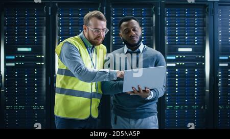 Bearded IT Specialist in Glasses and High Visibility Vest and Black Technician Colleague with a Laptop Computer Talking in Data Center while Standing Stock Photo