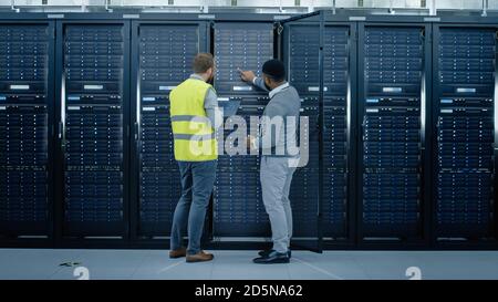 Bearded IT Engineer in Glasses and High Visibility Vest with a Laptop Computer and Black Technician Colleague Talking in Data Center while Standing Stock Photo