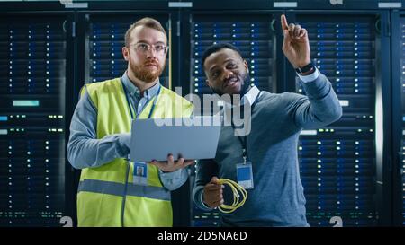 IT Specialist in Glasses and High Visibility Vest with Laptop Computer and Black Technician Colleague Talking in Data Center while Standing Next to Stock Photo