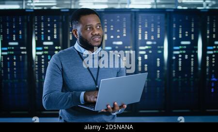 Black Data Center IT Technician Standing in the Middle of a Server Rack Corridor with a Laptop Computer. He is Visually Inspecting Working Server Stock Photo