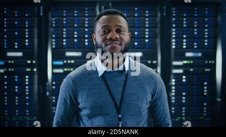 Bearded Black IT Engineer Standing and Smiling to the Camera in the Middle of a Working Data Center Server Room with Server Computers Working on a Stock Photo