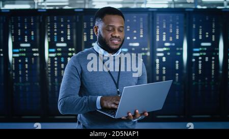 Black Data Center IT Technician Standing in the Middle of a Server Rack Corridor with a Laptop Computer. He is Visually Inspecting Working Server Stock Photo