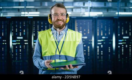Bearded IT Specialist in Glasses and Headphones, wearing High Visibility Vest is Smiles to the Camera in Data Center Next to Server Racks. He is Stock Photo