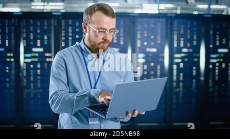 Bearded IT Specialist in Glasses is Working on Laptop in Data Center Next to Server Racks. Running Diagnostics or Doing Maintenance Work. Stock Photo