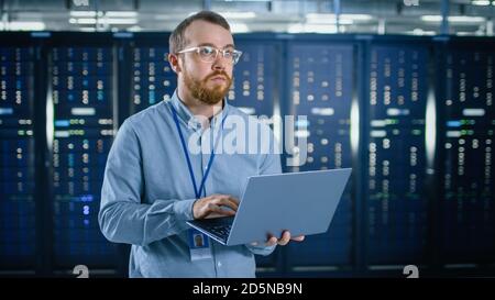 Bearded IT Specialist in Glasses is Working on Laptop in Data Center Next to Server Racks. Running Diagnostics or Doing Maintenance Work. Stock Photo