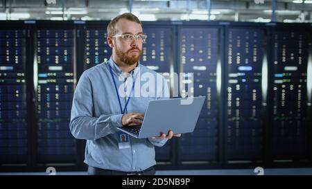 Bearded IT Specialist in Glasses is Working on Laptop in Data Center Next to Server Racks. Running Diagnostics or Doing Maintenance Work. Stock Photo