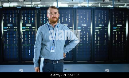 Bearded IT Specialist in Glasses is Standing in Data Center Next to Server Racks, Smiling to the Camera and Holding Laptop. Running Diagnostics or Stock Photo