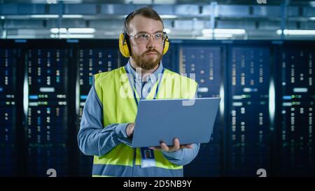 Bearded IT Specialist in Glasses and Headphones, wearing High Visibility Vest is Working on Laptop in Data Center Next to Server Racks. Stock Photo