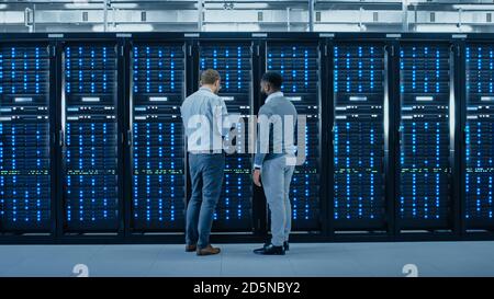Bearded IT Engineer in Glasses with a Laptop Computer and Black Technician Colleague Talking in Data Center while Standing Next to Server Racks Stock Photo