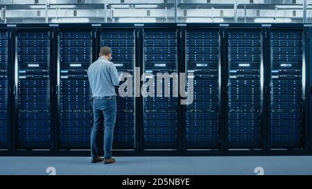 Bearded IT Specialist in Glasses is Working on Laptop in Data Center while Standing Before Server Rack. Running Diagnostics or Doing Maintenance Work. Stock Photo