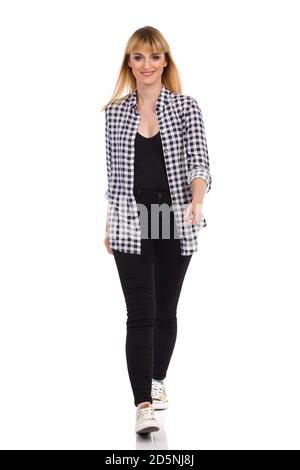 Confident young woman in unbuttoned lumberjack shirt, black jeans and sneakers is walking towards camera and smiling. Full length studio shot isolated Stock Photo