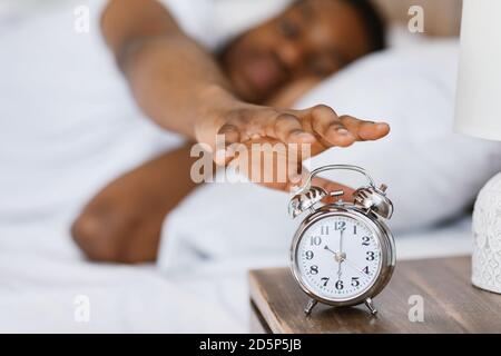 African Guy Waking Up Turning Off Alarm-Clock Lying In Bed Stock Photo