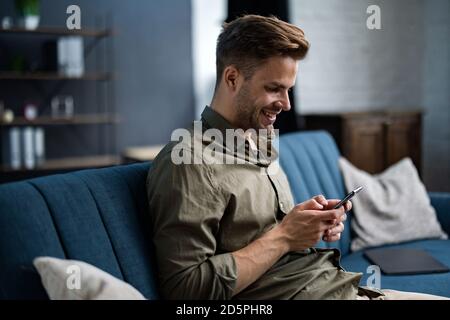 Young freelancer using smartphone and smiling. Happy man using mobile phone apps, texting message, browsing internet, looking at smartphone, sitting Stock Photo