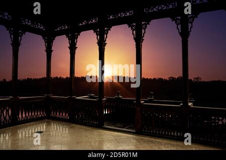 View of sunset on the lake in spring from wooden openwork gallery or gazebo. Forest and lake in the rays of the sun. Stock Photo
