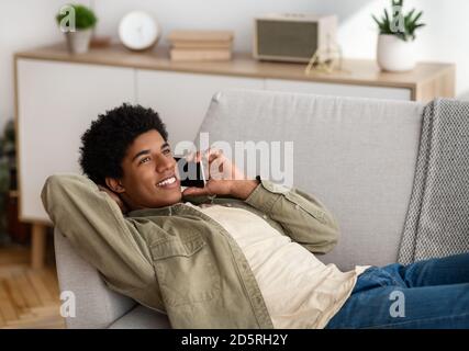 Funky African American teenager speaking on smartphone while lying on sofa at home, copy space Stock Photo