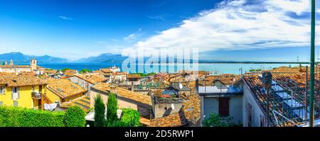 Panorama of Desenzano del Garda town with red tiled roof buildings, Garda Lake water, Monte Baldo mountain range, Sirmione peninsula, Lombardy, Northern Italy. Aerial panoramic view of Desenzano Stock Photo