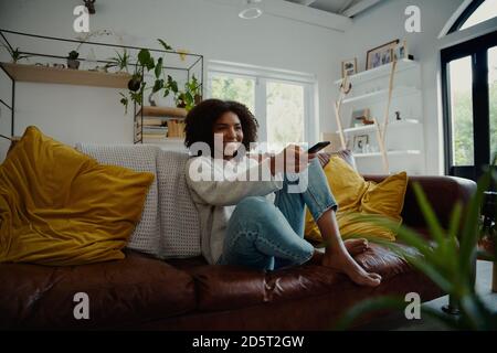 Beautiful young woman watching television sitting surrounded by cushions on a comfortable sofa with the remote control in her hand Stock Photo
