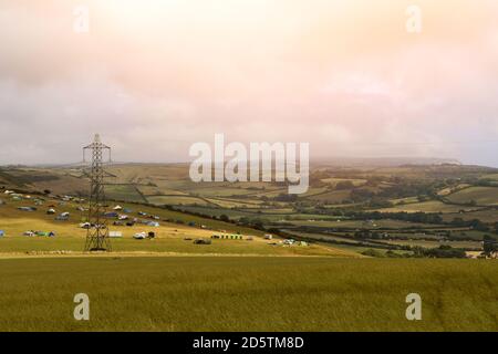 A view over the Dorset countryside with a camp site of tents in the distance and a lattice electricity tower . Stock Photo