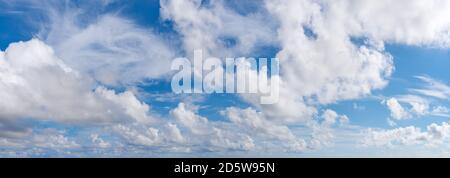 Fluffy cumulus clouds, translucent cirrus and stratus clouds float slowly across the bright blue sky on a sunny day. Panoramic skyscape shot. Weather. Stock Photo
