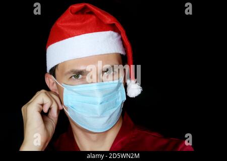 Portrait of man in medical face mask and Santa Claus hat on black background. Christmas celebration during coronavirus pandemic Stock Photo