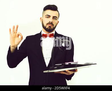 Work done successfully. OK sign shown by successful employer in suit and red bow, holding office appliances with laptop and desk lamp, isolated on white background. Concept of business and office work Stock Photo